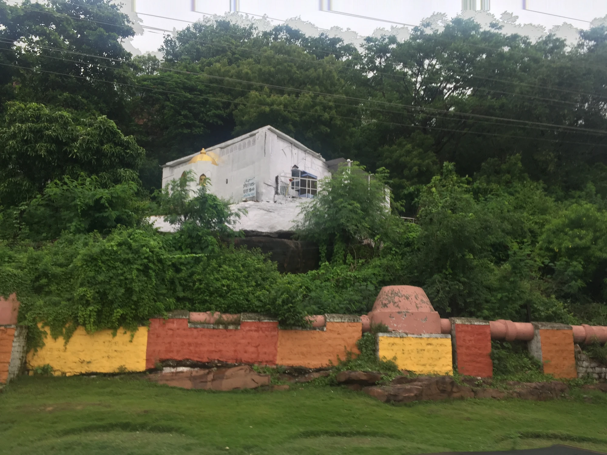 A white shrine on a rock outcrop surrounded by trees, seen over a painted boundary wall