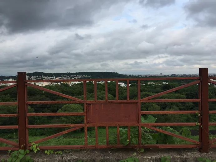 A rusty metal gate overlooking a forested cityscape under monsoon clouds