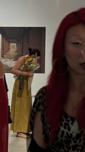 A woman in a yellow dress leaning over flowers, with a red-haired woman in the foreground