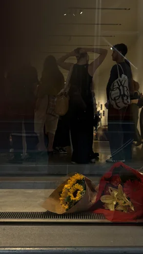 Reflections of figures in a gallery window above a bunch of yellow and red flowers