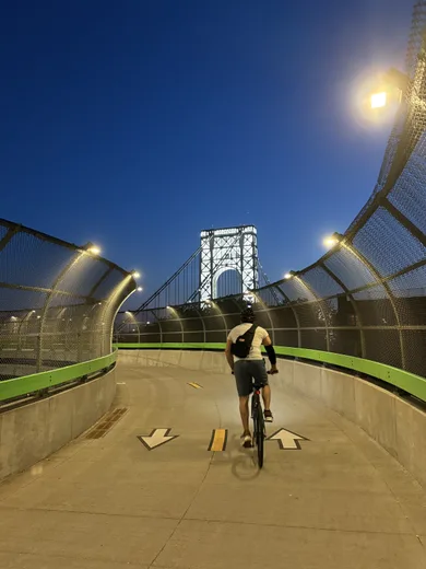 A cyclist on a lit riverside bridge path with the George Washington Bridge above